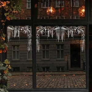 Peut inclure: Un autocollant décoratif blanc pour fenêtre présentant un motif botanique symétrique avec des guirlandes florales suspendues, des feuilles délicates et des lignes pointillées. Le motif complexe est appliqué sur deux vitres sombres.