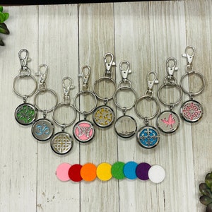 May include: A collection of silver keychains with various decorative pendants, including a tree, Om symbol, butterfly, and geometric designs. Colorful felt pads are arranged below. The keychains have a silver clasp and ring.