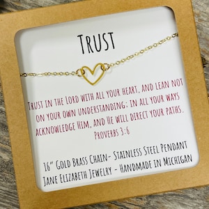 May include: A gold-colored necklace with a heart-shaped pendant, displayed on a white card inside a brown box. The card features the word "TRUST" and a Bible verse. The necklace is 16 inches long.