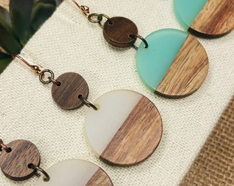 May include: Three pairs of earrings with a wood and resin design. Each earring features a round wooden disc with a half-circle of resin on top. The resin is in three different colours: white, light blue, and a darker blue.