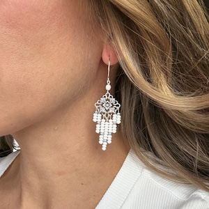 May include: Silver chandelier earrings with a floral filigree design and dangling white beads. The earrings have a hook closure and are shown on a person's ear. The earrings are a classic accessory.