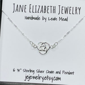 May include: A silver chain necklace with a Celtic knot pendant. The necklace is 16 inches long and is made of sterling silver. The pendant is a classic Celtic knot design. The necklace is displayed in a white box with the text "JANE ELIZABETH JEWELRY Handmade by Leah Mead 16 34" Sterling Silver Chain and Pendant jejewelry.etsy.com".