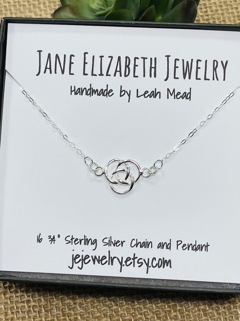 May include: A silver chain necklace with a Celtic knot pendant. The necklace is 16 inches long and is made of sterling silver. The pendant is a classic Celtic knot design. The necklace is displayed in a white box with the text "JANE ELIZABETH JEWELRY Handmade by Leah Mead 16 34" Sterling Silver Chain and Pendant jejewelry.etsy.com".