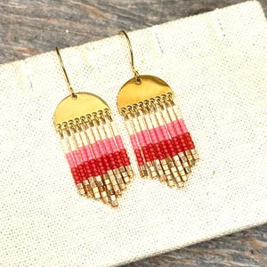 May include: Gold-coloured earrings with a half-circle top and beaded fringe. The fringe has alternating rows of white, pink, and red beads, with gold square beads at the bottom. The earrings are displayed on a white fabric.