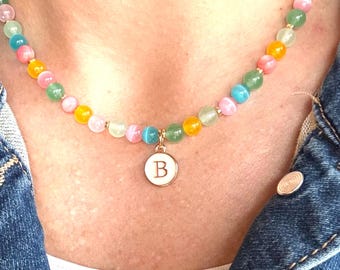 May include: A colorful beaded necklace with a pendant featuring the letter "B". The necklace has a variety of pastel-colored beads, including pink, yellow, green, and blue. The pendant is white with a gold-colored border.