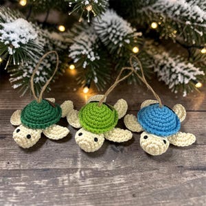May include: Three crocheted sea turtle ornaments with cream-colored bodies and shells in dark green, lime green, and blue. Each turtle features small black bead eyes and a brown twine loop attached to the top of the shell for hanging purposes.