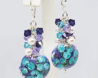 Peut inclure: Boucles d'oreilles pendantes florales en turquoise et violet avec des accents de perles et de cristaux. Les boucles d'oreilles sont faites à la main et présentent un design floral unique.