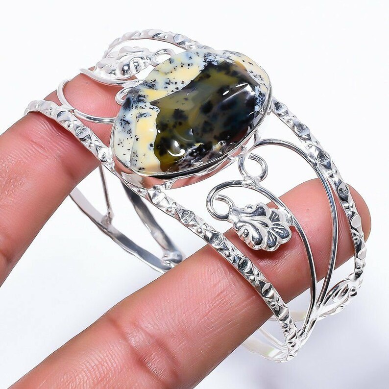 Peut inclure: Un bracelet manchette en argent avec une grande pierre d'opale de forme ovale. L'opale est un m&eacute;lange de couleurs blanches, noires et vertes. Le bracelet pr&eacute;sente un motif floral d&eacute;licat.