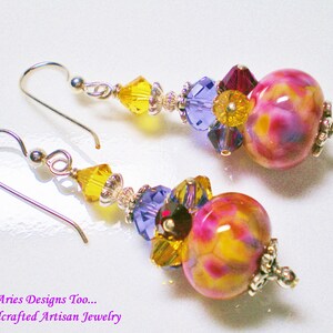 May include: A pair of dangle earrings with yellow, purple, and pink glass beads. The earrings have silver hooks and are accented with clear and purple crystals.