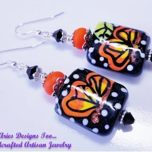 May include: A pair of dangle earrings with black, orange and white square beads. The beads feature a butterfly design with black outlines and white polka dots. The earrings are made with silver-toned metal.