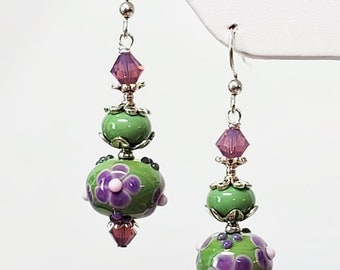 以下が含まれることがあります： 手作りの職人技のイヤリングのペア。各イヤリングは、紫色の花のアクセントが付いた緑色のガラスビーズ、小さな緑色のビーズ、紫色のクリスタルが特徴です。イヤリングは、シルバートーンのイヤーフックから吊り下げられています。