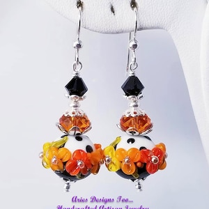 Pode incluir: Um par de brincos pendentes com uma conta de bolinhas pretas e brancas, contas de flores laranja e amarelas e uma conta de cristal preto no topo. Os brincos estão em um expositor branco. "Aries Designs Too... Handcrafted Artisan Jewelry" está escrito abaixo dos brincos.