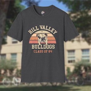 May include: Dark grey t-shirt with a retro design. The shirt features the text "HILL VALLEY BULLDOGS CLASS OF 84" above a bulldog illustration set against a sunset-style background. The t-shirt has a crew neck and short sleeves.