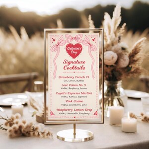May include: A gold-framed sign with a light background displays "Galentine's Day Signature Cocktails." The menu lists cocktails like Strawberry French 75 and Love Potion No. 9. The sign is on a gold stand, with candles and floral arrangements in the background.