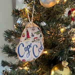 May include: A decorative oyster shell ornament featuring a hand-painted design of lighthouses, sailboats, and seashells with the text Cape Cod written in blue script. The shell has a gold-painted edge and hangs from a thin white twine string.