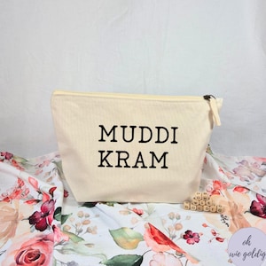 Könnte beinhalten: Eine cremefarbene Canvas-Tasche mit Reißverschluss und dem schwarzen Aufdruck "MUDDI KRAM". Die Tasche liegt auf einem Stoff mit Blumenmuster in Rot, Rosa und Grün.