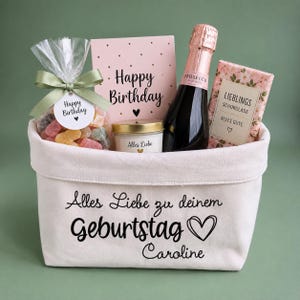 Könnte beinhalten: Ein cremefarbener Stoffkorb mit dem Aufdruck Alles Liebe zu deinem Geburtstag Caroline und einem Herz. Darin befinden sich eine Flasche Prosecco, eine rosa Geburtstagskarte, eine Kerze mit der Aufschrift Alles Liebe, eine Schokolade und bunte Bonbons.