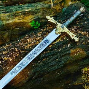 Puede incluir: Una espada larga y plateada con empu&ntilde;adura y guarda doradas, apoyada sobre un tronco desgastado. La hoja tiene un dise&ntilde;o grabado. La espada es un art&iacute;culo de fantas&iacute;a o hist&oacute;rico.