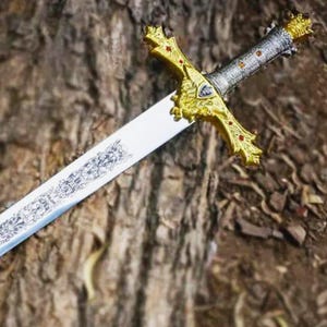 May include: A decorative sword with a silver blade and a gold-colored hilt. The hilt features intricate designs and red accents. The sword is lying on a textured, brown surface, possibly wood.