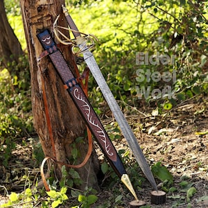 Op de afbeelding: Een rapier met een patroon in het stalen lemmet, een messing gevest en een geribbelde greep. De bijbehorende zwarte lederen schede heeft een bruine lederen overlay met een wit rankenpatroon, een messing punt en lederen riemen. Tekst Elder Steel Work.