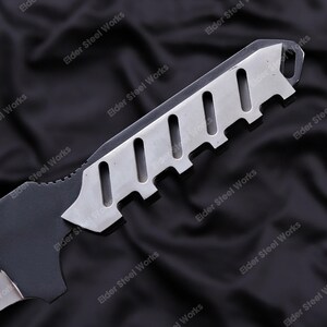 May include: Close-up of a black and silver knife. The blade is black with a serrated edge. The handle is silver with a series of rectangular cutouts. The knife is set against a dark background.