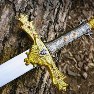 Puede incluir: Una espada decorativa con una hoja plateada y una empu&ntilde;adura dorada. La empu&ntilde;adura est&aacute; adornada con piedras preciosas rojas y naranjas. La espada descansa sobre una superficie marr&oacute;n texturizada, posiblemente un tronco de &aacute;rbol.