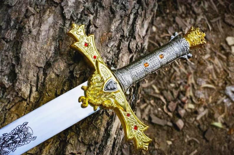 Puede incluir: Una espada decorativa con una hoja plateada y una empu&ntilde;adura dorada. La empu&ntilde;adura est&aacute; adornada con piedras preciosas rojas y naranjas. La espada descansa sobre una superficie marr&oacute;n texturizada, posiblemente un tronco de &aacute;rbol.