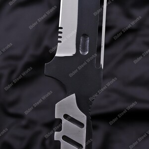 May include: Close-up of a black and silver knife blade. The blade has a serrated edge and a series of cutouts. The text "Elder Steel Works" is printed on the blade.