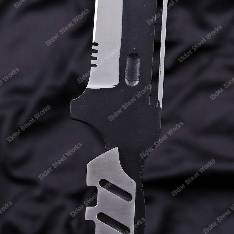 May include: Close-up of a black and silver knife blade. The blade has a serrated edge and a series of cutouts. The text "Elder Steel Works" is printed on the blade.