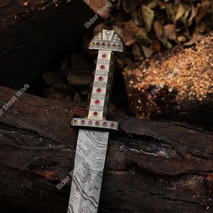 May include: A decorative sword with a patterned blade and an ornate handle. The handle is silver and gold with red accents. The sword is resting on a bed of dark brown wood.