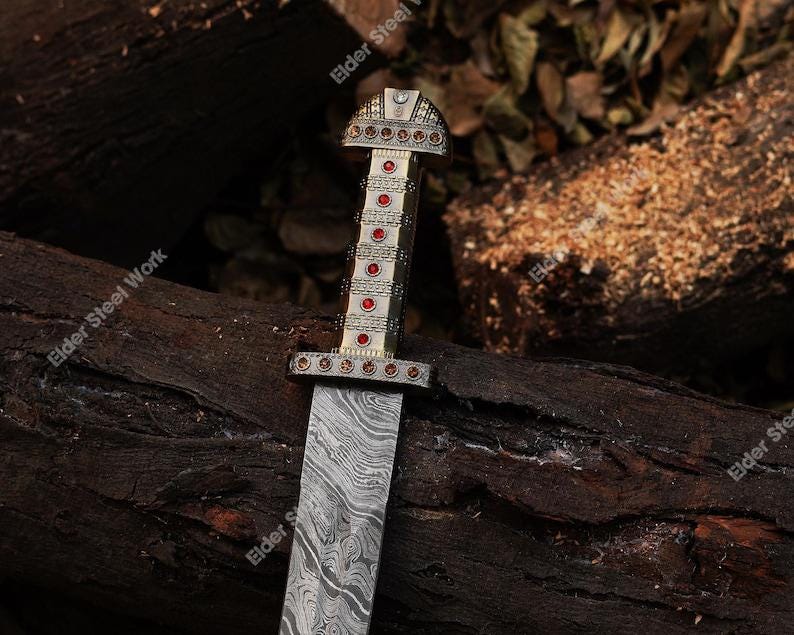 May include: A decorative sword with a patterned blade and an ornate handle. The handle is silver and gold with red accents. The sword is resting on a bed of dark brown wood.
