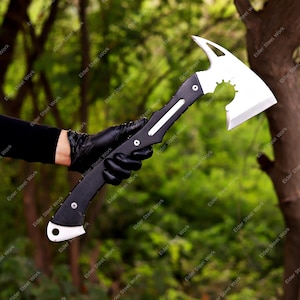 Könnte beinhalten: Eine taktische Axt mit schwarzem Griff und silberner Klinge. Der Axtkopf hat ein einzigartiges Design mit einer gebogenen Klinge und einem gezahnten Abschnitt. Der Griff wird von einer behandschuhten Hand vor einem verschwommenen grünen Hintergrund gehalten.