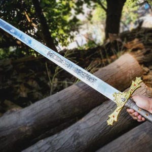 Puede incluir: Una espada larga y ornamentada con una hoja plateada y una empu&ntilde;adura dorada. La empu&ntilde;adura presenta detalles rojos y un dise&ntilde;o detallado. La espada se sostiene sobre un fondo de troncos de madera y vegetaci&oacute;n.