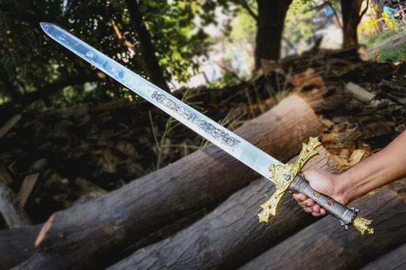 Puede incluir: Una espada larga y ornamentada con una hoja plateada y una empu&ntilde;adura dorada. La empu&ntilde;adura presenta detalles rojos y un dise&ntilde;o detallado. La espada se sostiene sobre un fondo de troncos de madera y vegetaci&oacute;n.