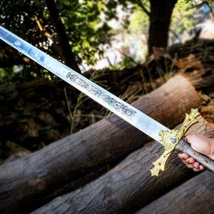 Puede incluir: Una espada larga y plateada con una empu&ntilde;adura dorada ornamentada y un dise&ntilde;o detallado en la hoja. La espada se sostiene sobre un fondo de troncos y follaje, mostrando su artesan&iacute;a y est&eacute;tica hist&oacute;rica.