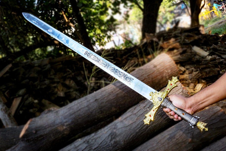 Puede incluir: Una espada larga y plateada con una empu&ntilde;adura dorada ornamentada y un dise&ntilde;o detallado en la hoja. La espada se sostiene sobre un fondo de troncos y follaje, mostrando su artesan&iacute;a y est&eacute;tica hist&oacute;rica.