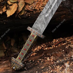 May include: Close-up of a decorative sword with a patterned blade and ornate handle. The handle is gold-toned with red and clear jewel accents. The blade has a unique, swirling pattern. The sword is set against a natural, wood-like background.
