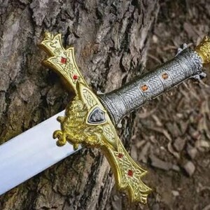 Puede incluir: Una espada decorativa con una hoja plateada y una empu&ntilde;adura dorada ornamentada. La empu&ntilde;adura presenta detalles rojos y un dise&ntilde;o detallado. La espada descansa sobre una superficie marr&oacute;n texturizada, sugiriendo un tema hist&oacute;rico o de fantas&iacute;a.