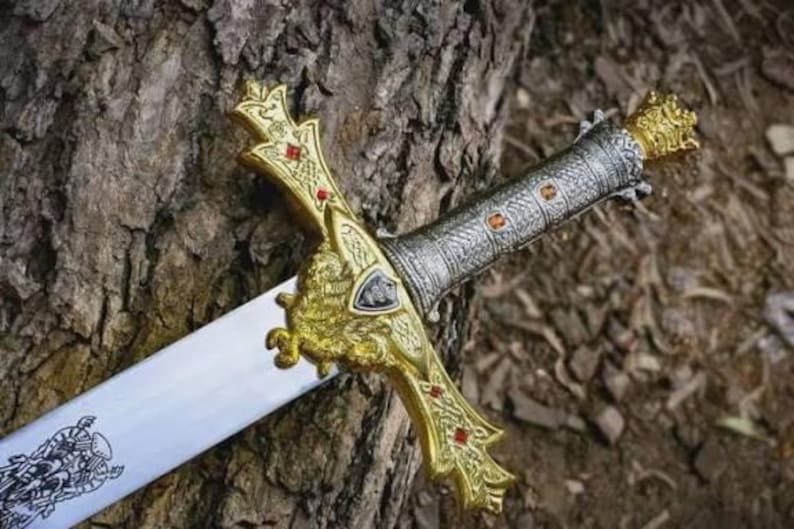 Puede incluir: Una espada decorativa con una hoja plateada y una empu&ntilde;adura dorada ornamentada. La empu&ntilde;adura presenta detalles rojos y un dise&ntilde;o detallado. La espada descansa sobre una superficie marr&oacute;n texturizada, sugiriendo un tema hist&oacute;rico o de fantas&iacute;a.