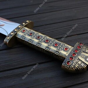 May include: A close-up of a decorative sword with a bronze-colored handle and a silver blade. The handle is adorned with red gemstones and intricate metalwork. The sword rests on a dark wooden surface.