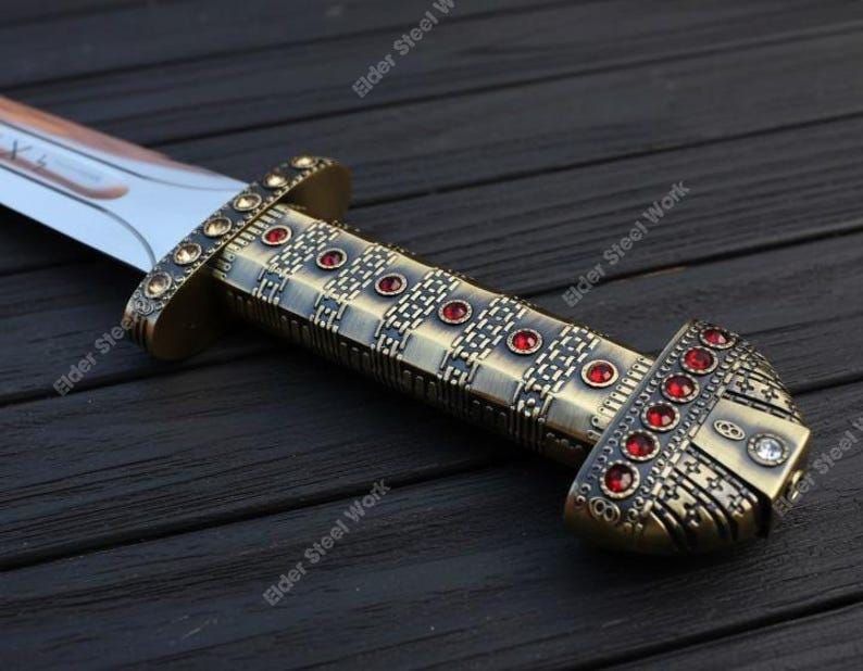 May include: A close-up of a decorative sword with a bronze-colored handle and a silver blade. The handle is adorned with red gemstones and intricate metalwork. The sword rests on a dark wooden surface.