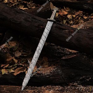 May include: A long, silver sword with a patterned blade and ornate handle rests on a dark wooden surface. The blade has a wavy design, and the handle features intricate detailing. The sword is a historical weapon.