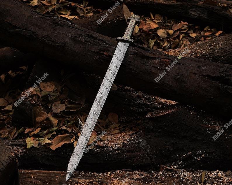 May include: A long, silver sword with a patterned blade and ornate handle rests on a dark wooden surface. The blade has a wavy design, and the handle features intricate detailing. The sword is a historical weapon.