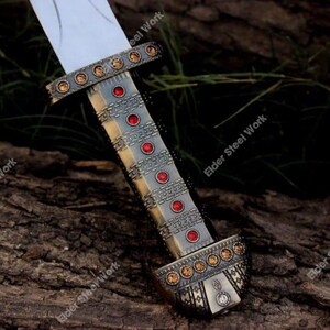 May include: A close-up of a decorative sword with a silver blade and ornate handle. The handle is detailed with red and gold accents, and the text "Elder Steel Work" is visible.