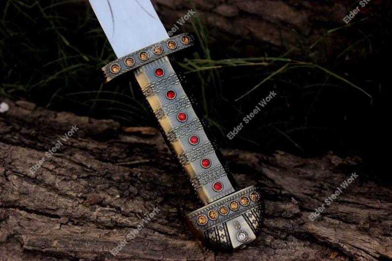 May include: A close-up of a decorative sword with a silver blade and ornate handle. The handle is detailed with red and gold accents, and the text "Elder Steel Work" is visible.