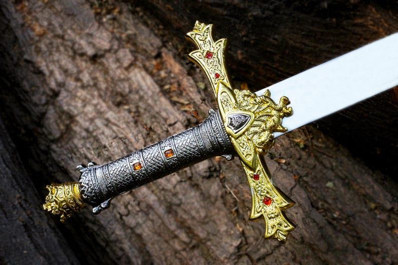 Puede incluir: Primer plano de una espada decorativa. La empu&ntilde;adura es plateada con detalles dorados y joyas naranjas. La guarda es dorada con joyas rojas. La hoja blanca se apoya sobre un fondo marr&oacute;n texturizado.