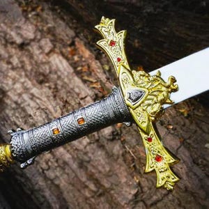 Puede incluir: Una espada decorativa con una hoja blanca y una empu&ntilde;adura ornamentada en oro y plata. La empu&ntilde;adura presenta joyas rojas y un dise&ntilde;o detallado. La espada se encuentra sobre un fondo de madera texturizada.