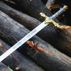 Puede incluir: Una espada decorativa con una hoja plateada y una empu&ntilde;adura dorada ornamentada. La empu&ntilde;adura presenta un dise&ntilde;o de cruz con detalles rojos. La espada descansa sobre un fondo de troncos de madera apilados, creando una est&eacute;tica r&uacute;stica.