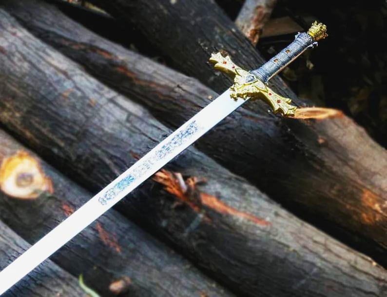 Puede incluir: Una espada decorativa con una hoja plateada y una empu&ntilde;adura dorada ornamentada. La empu&ntilde;adura presenta un dise&ntilde;o de cruz con detalles rojos. La espada descansa sobre un fondo de troncos de madera apilados, creando una est&eacute;tica r&uacute;stica.