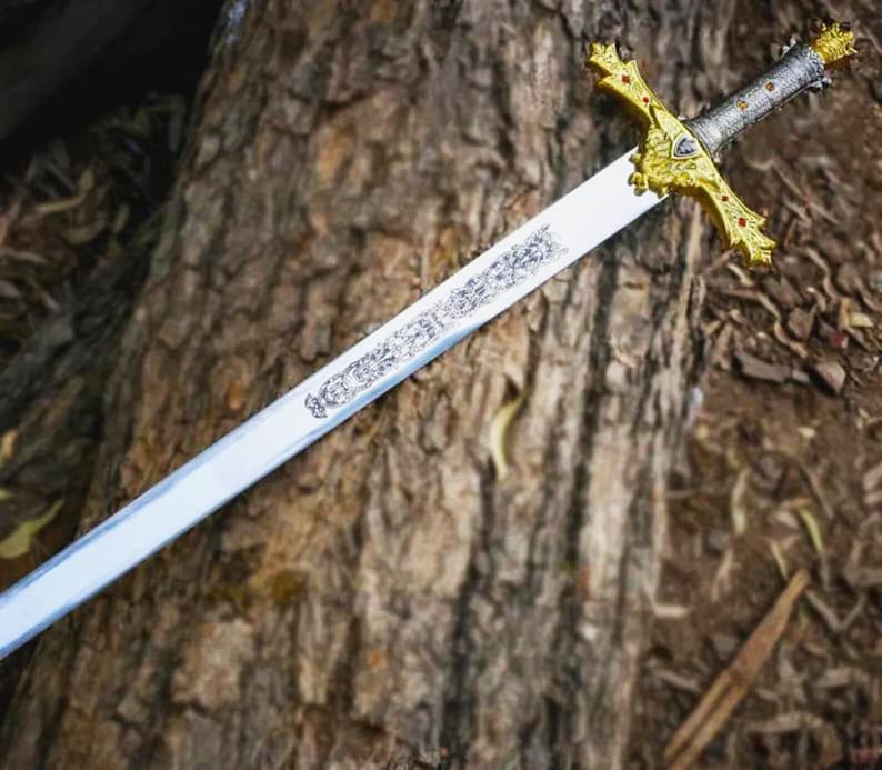 Puede incluir: Una espada decorativa con una hoja plateada y una empu&ntilde;adura dorada. La empu&ntilde;adura presenta dise&ntilde;os intrincados y detalles rojos. La espada est&aacute; sobre una superficie marr&oacute;n texturizada, posiblemente madera.